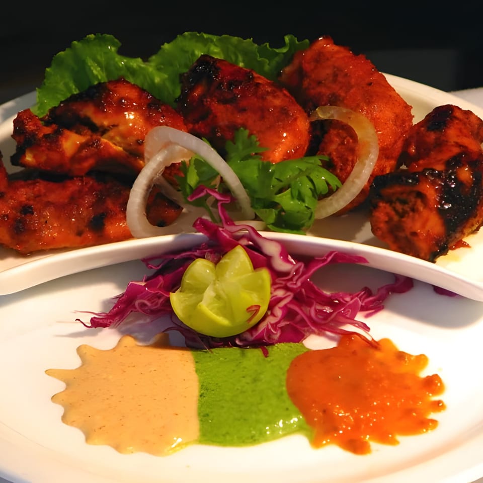 Indian chicken tikka with onions, lettuce, lime, red cabbage, and three sauces on a white plate.