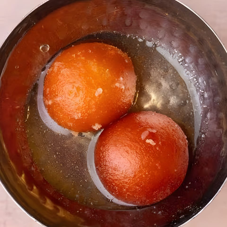 Two orange gulab jamun in a silver bowl, soaking in sweet syrup.