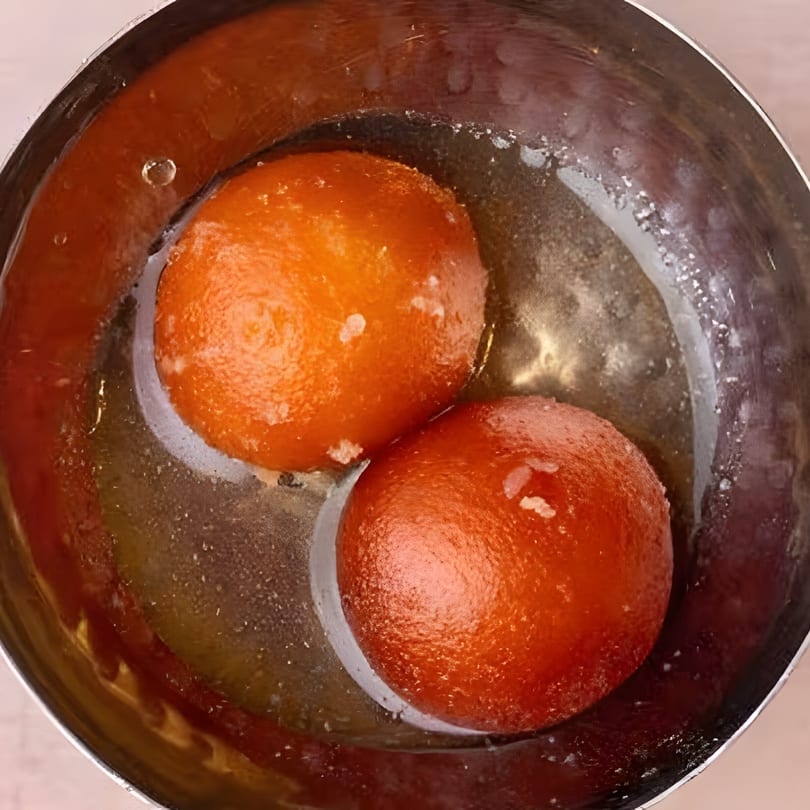 Two orange gulab jamun in a silver bowl, soaking in sweet syrup.
